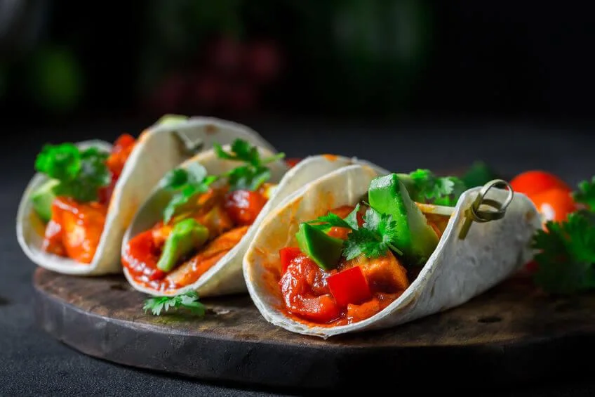 Tres tacos con palta y tomates cortados a la vista, posados sobre una tabla de madera.